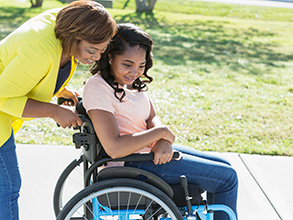 A smiling girl in a wheelchair while being pushed by her mother on a sidewalk